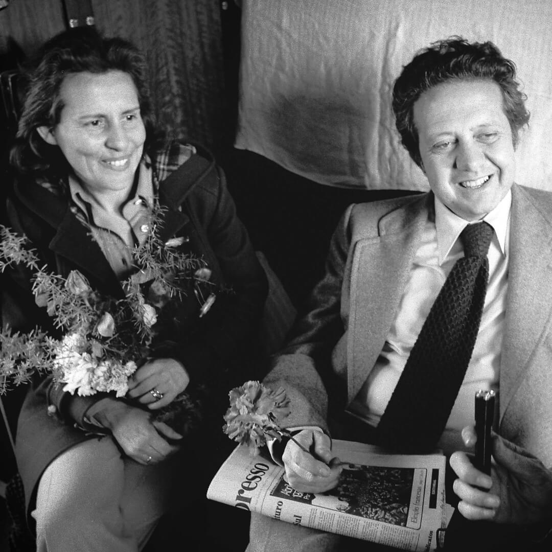 Fotografia a preto e branco de duas pessoas sentadas, uma segurando flores e a outra um jornal